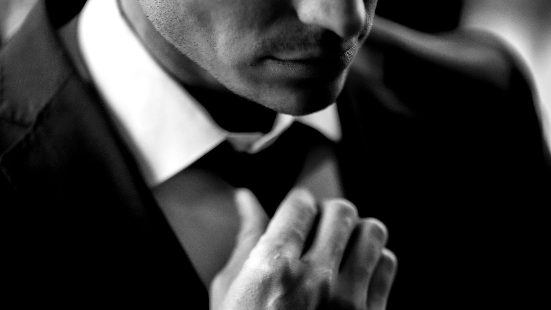 A close-up greyscale image of a suited man adjusting his tie