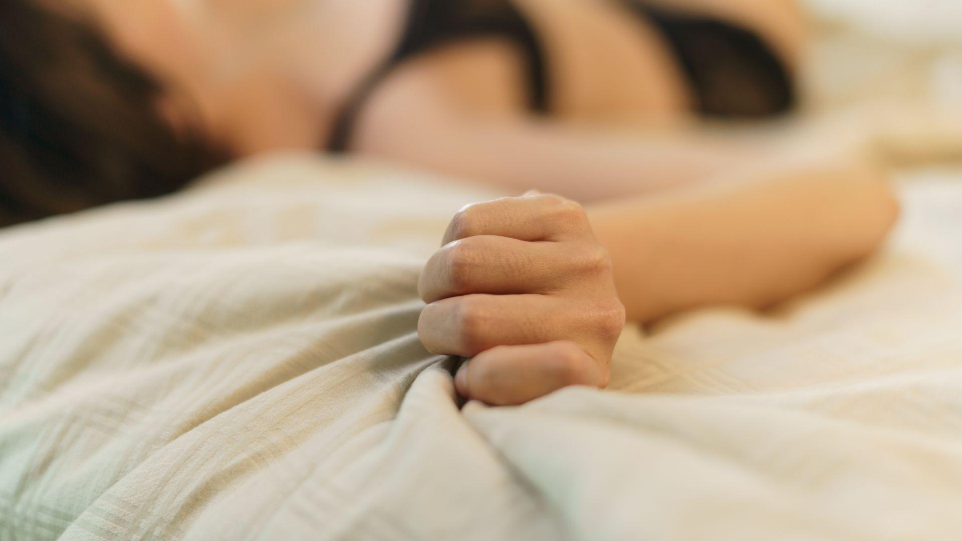 A woman grips the bedsheets, suggesting orgasm