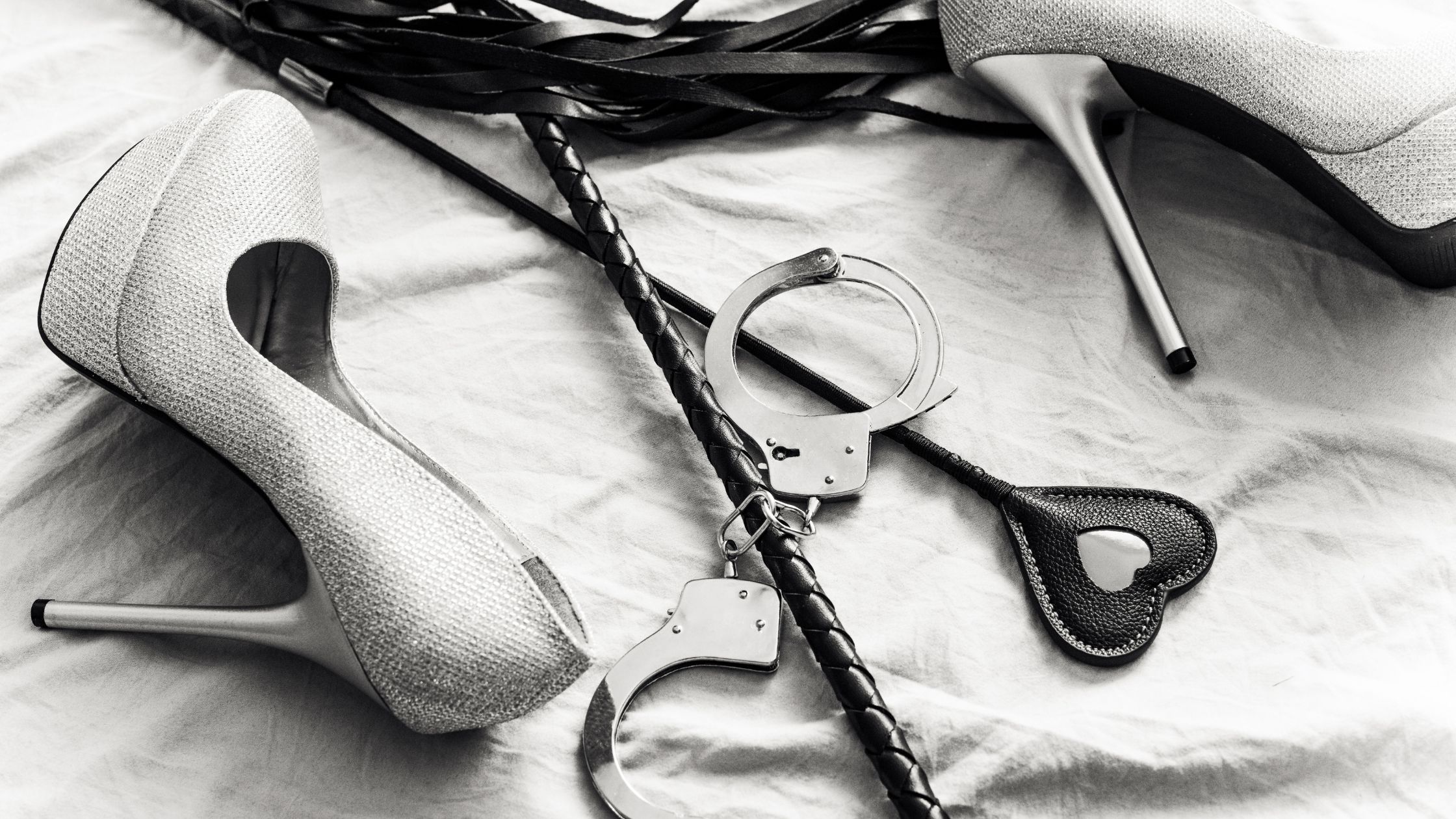 Lessons in BDSM header image showing black leather BDSM accessories and silver high heels on a white bedspread.