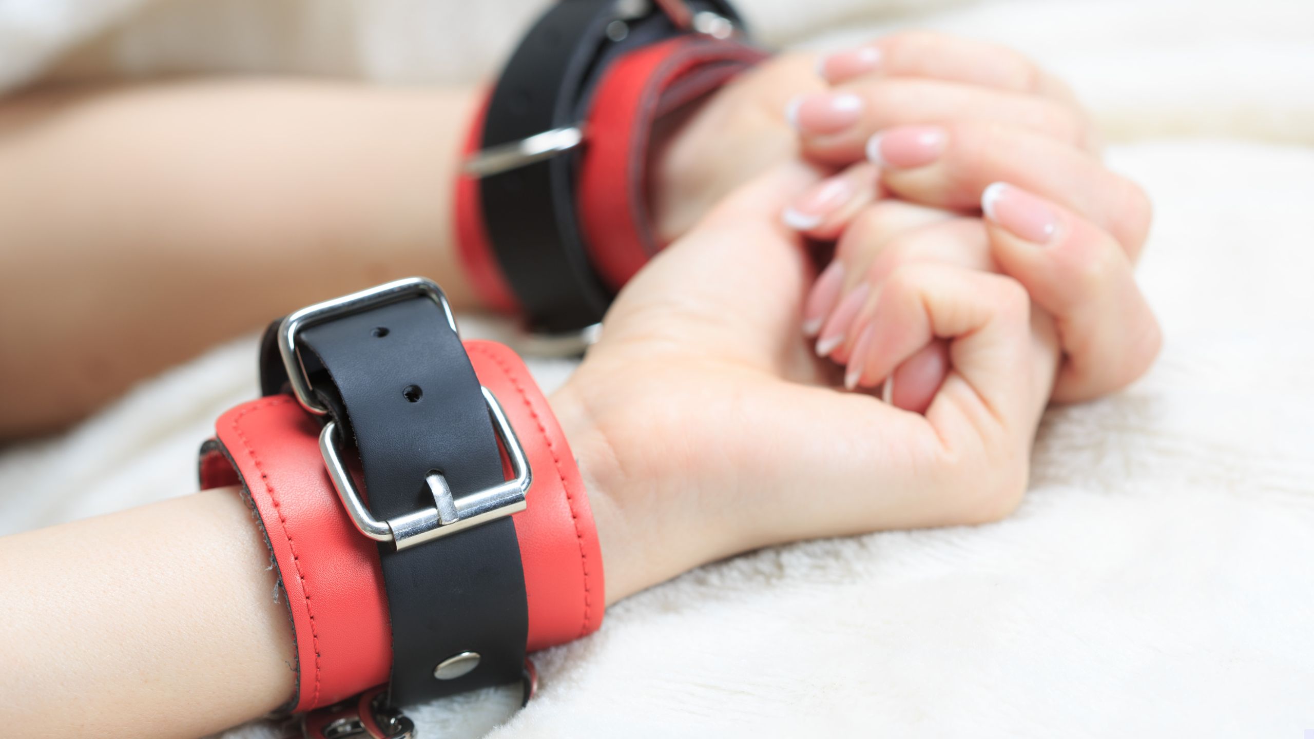 A woman's hands bound in red and black leather bondage cuffs on a fluffy white background
