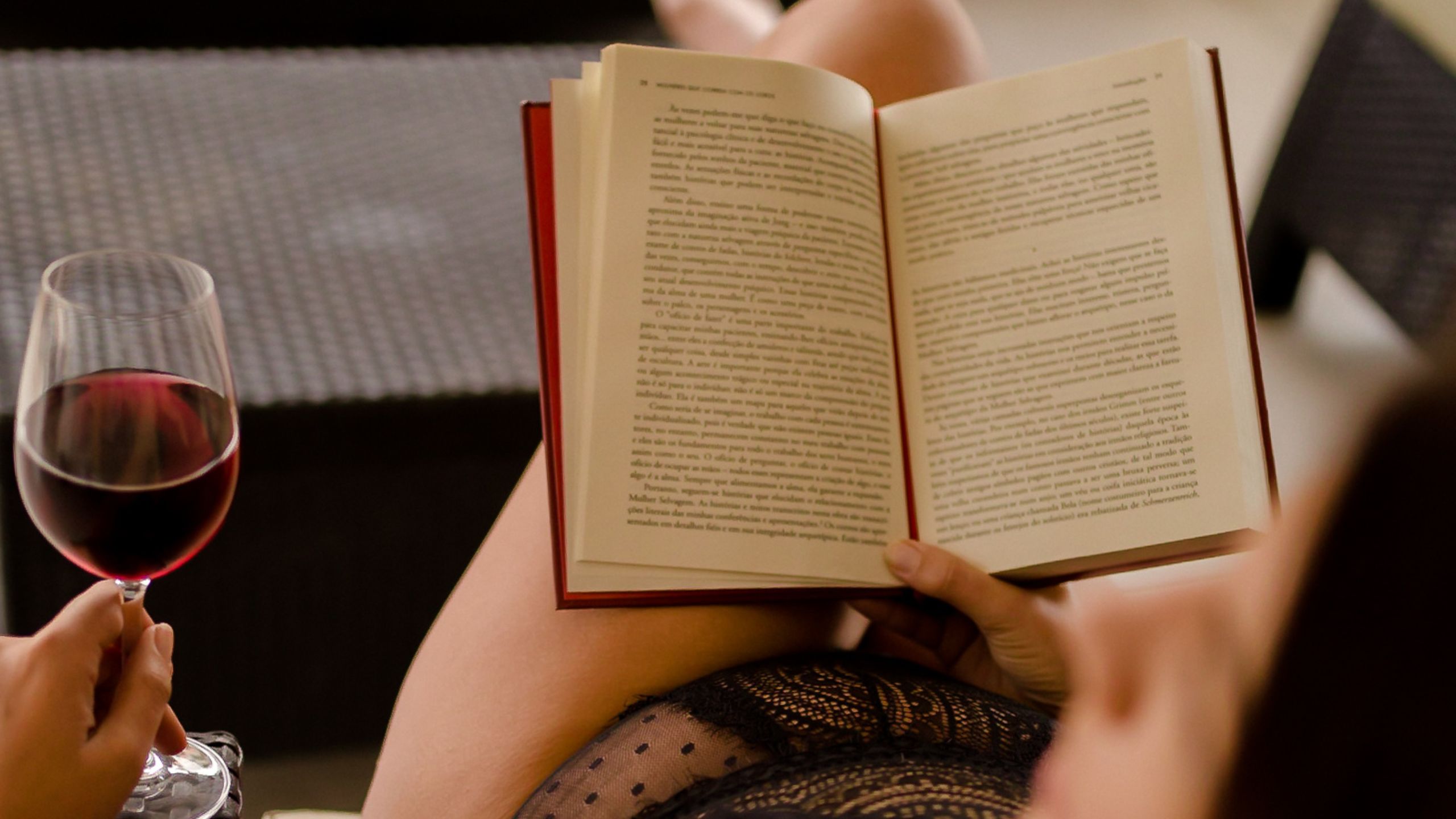 A sexy woman in black linger reads a book and holds a glass of red wine