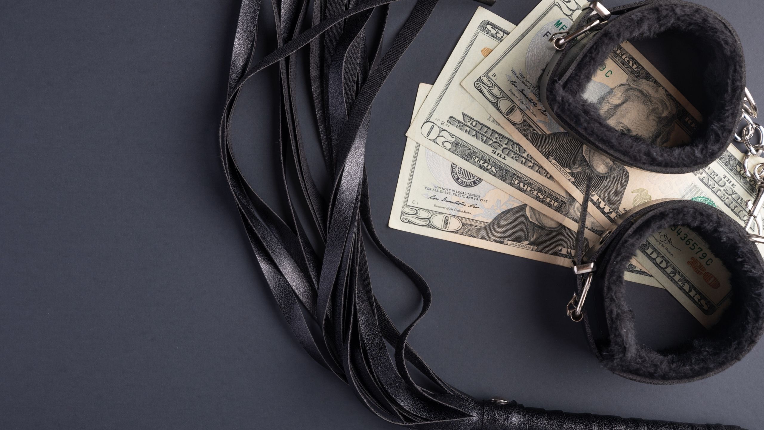 Black leather BDSM accessories, including cuffs and a flogger, arranged alongside US dollar bills on a dark background, illustrating kink on a budget.