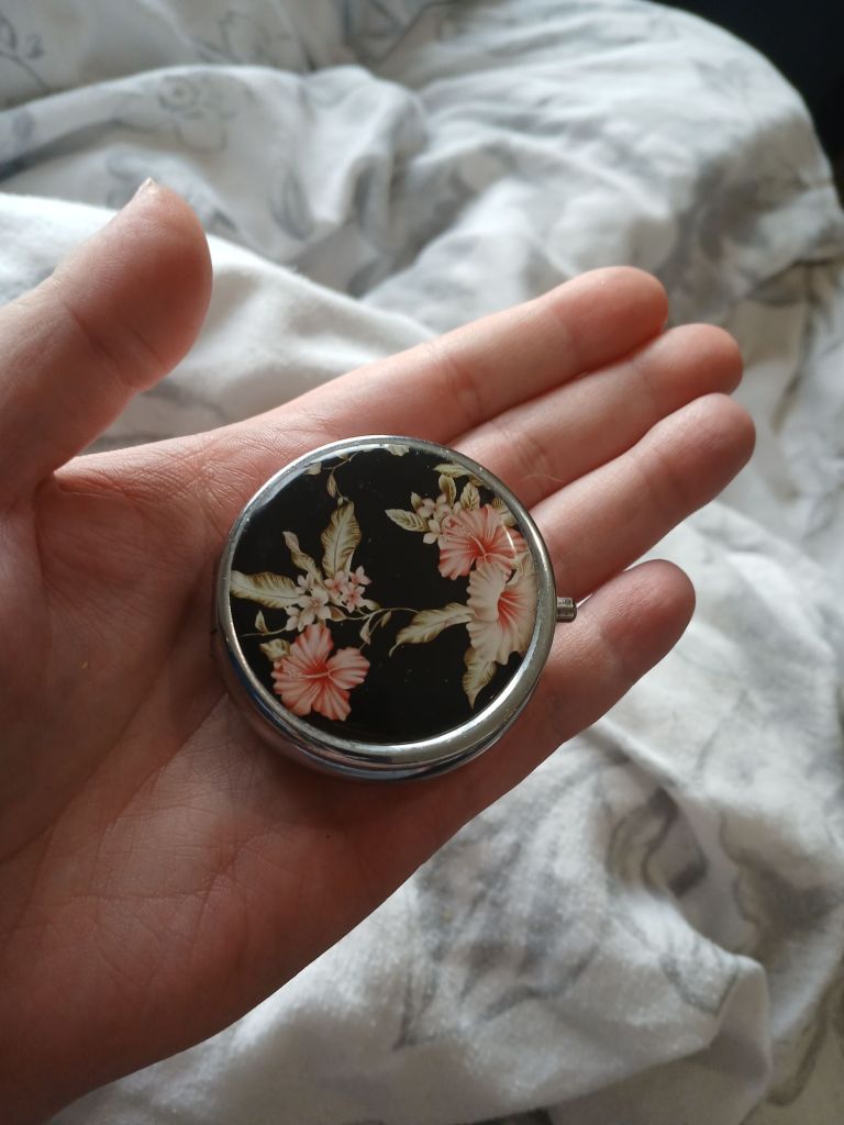 A floral pill pot held in the aythor's hand, over floral grey bedding