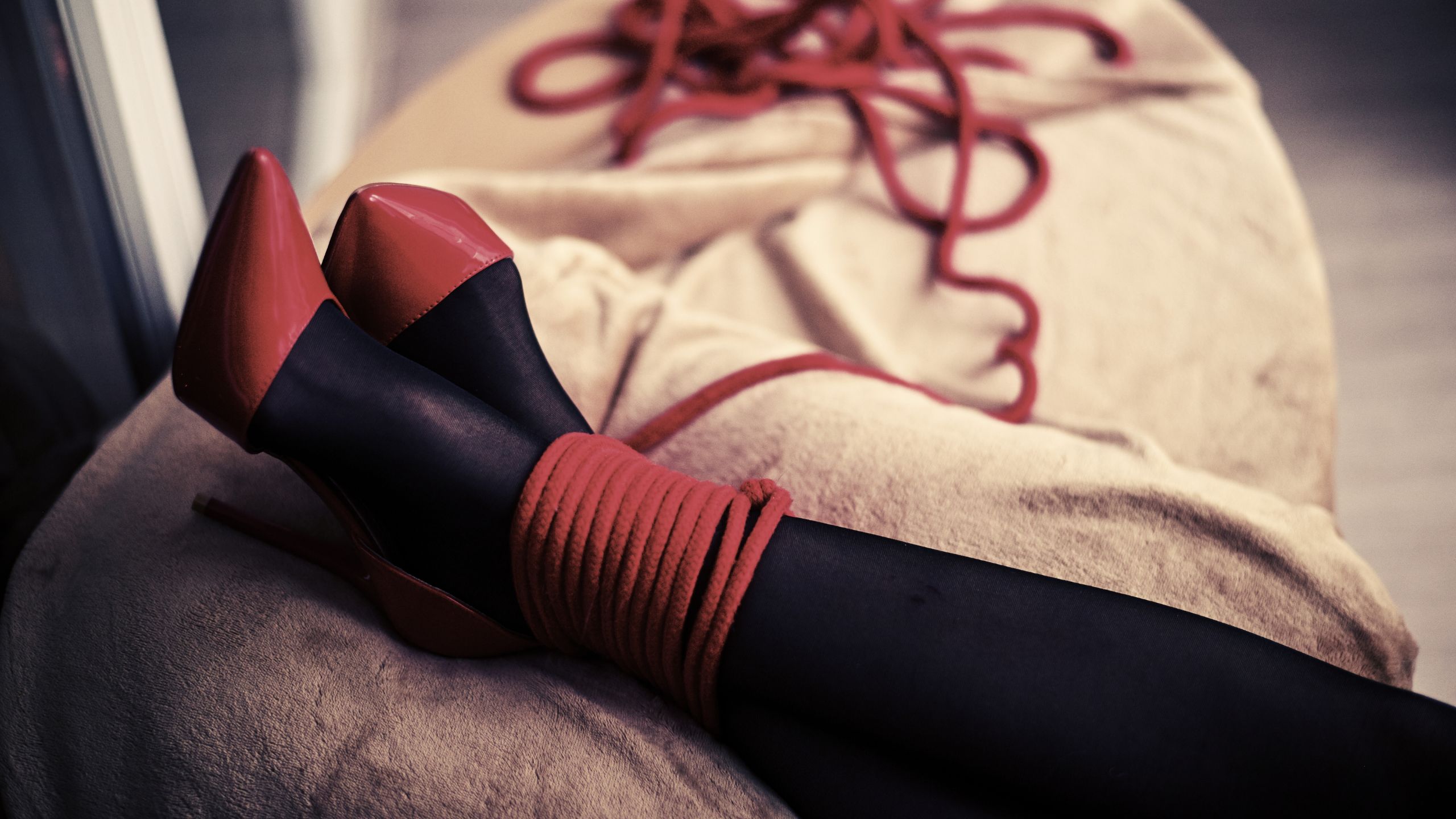 A person lying on a cream bed with their ankles crossed and bound in red rope. They are wearing black stockings and red high heels with more red rope beside them.